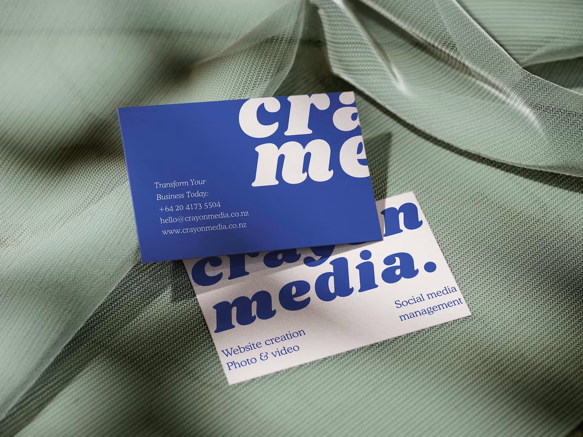 Two business cards on a textured green surface. The top card is dark blue with white text that includes "crayme" and contact details. The bottom card is white with blue text stating "crayme media" and services like "Website creation" and "Photo & video."