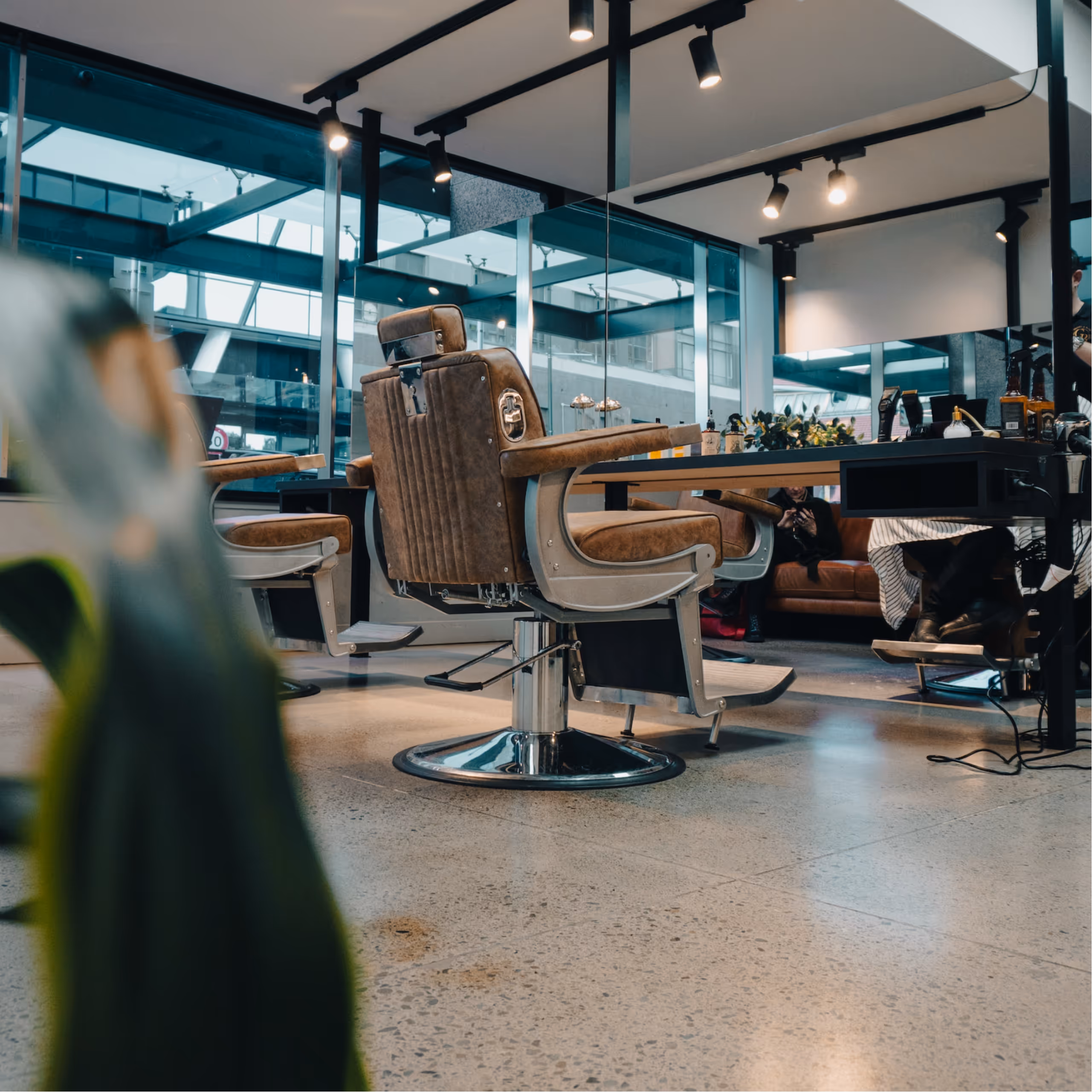 A modern barber shop interior featuring two brown leather barber chairs, a small side table, and large windows allowing natural light, with a comfortable couch in the corner.